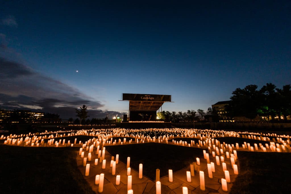 A wide shot of candles spread out across an outdoor area with a stage set up for an open-air Candlelight concert in the distance.