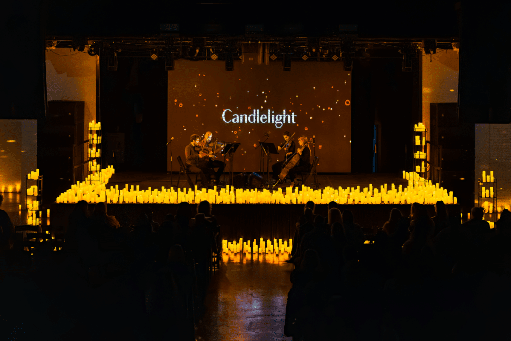 A string quartet performing on a raised stage surrounded by hundreds of candles with a big screen behind them displaying the word 